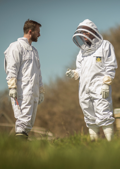 Two individuals wearing white apiarist suits with veils, standing outdoors.