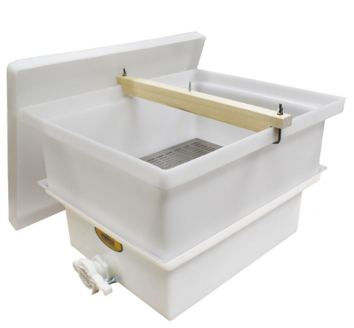 A white plastic honey straining tank used in beekeeping, shown with the lid removed, a wooden bar mounted across the top to hold frames, a metal strainer inside the basin, and a spigot on the front for draining filtered honey