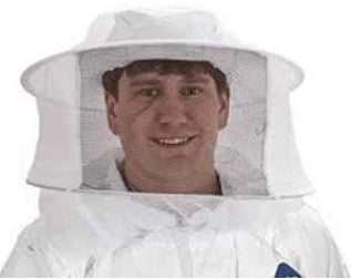 A person wearing a white beekeeping suit with a round mesh veil that covers the head and face. The veil is supported by a circular brim, creating space between the mesh and the wearer’s face. The person is smiling and shown from the shoulders up against a plain background.