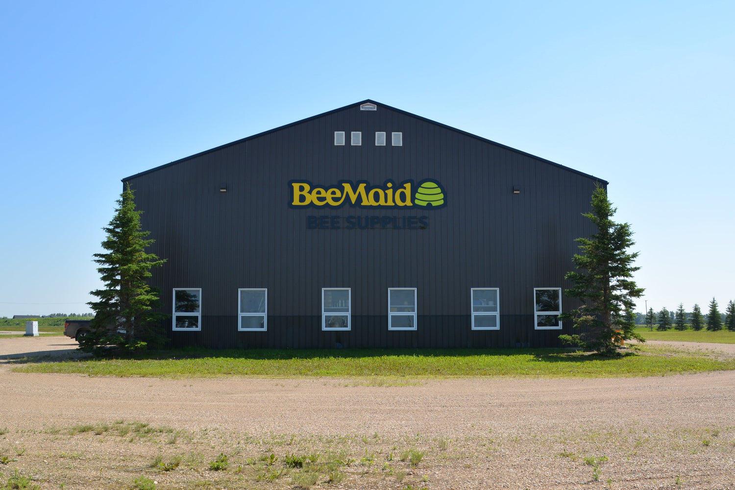 A large dark gray BeeMaid Bee Supplies building with yellow and green logo lettering, situated in an open grassy field under a clear blue sky.